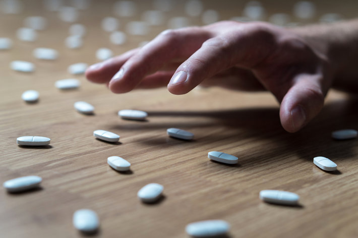A hand is reaching for pills which are laying all over the floor.